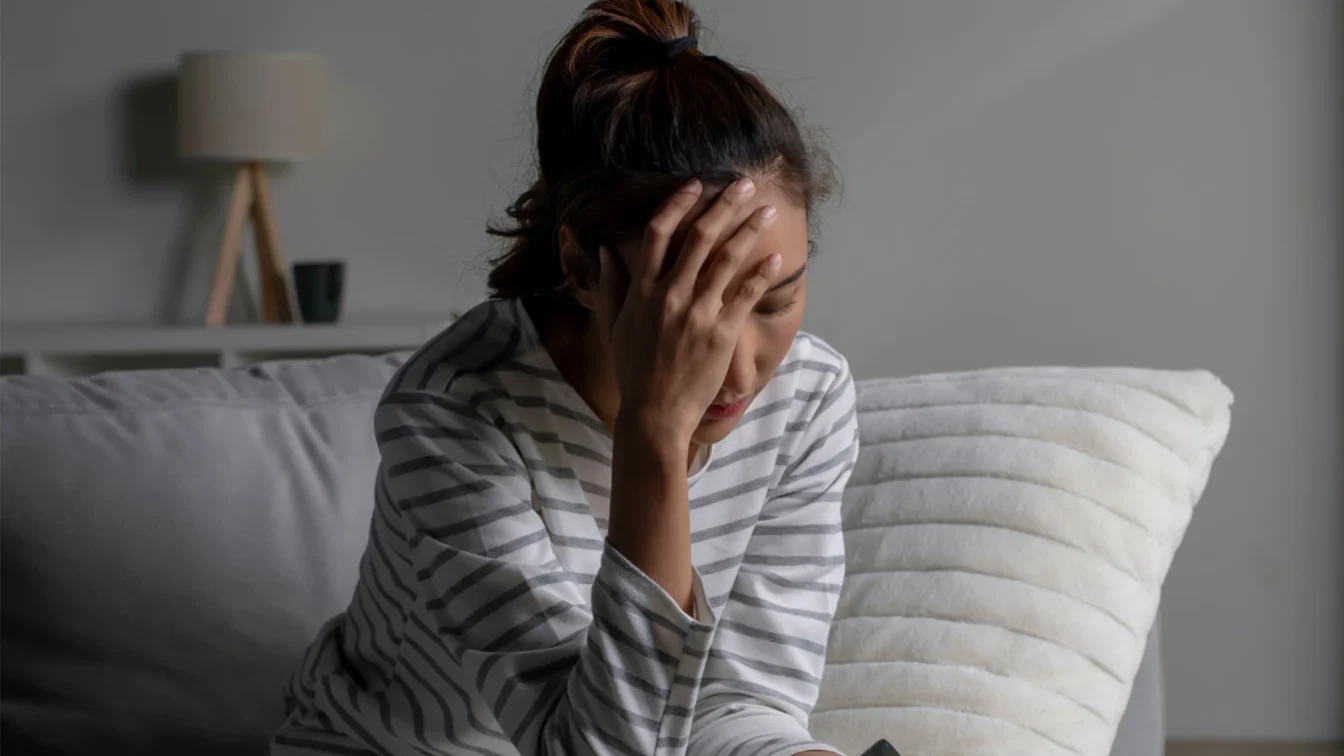 Imagen de una mujer sentada en un sofá mientras se toca la frente con la palma de la mano con evidentes síntomas de dolor y/o depresión