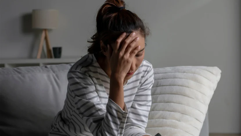 Imagen de una mujer sentada en un sofá mientras se toca la frente con la palma de la mano con evidentes síntomas de dolor y/o depresión